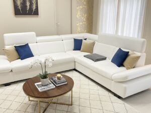 Canapé d'angle blanc moderne avec coussins bleus et dorés, table basse en bois et tapis à motifs géométriques.