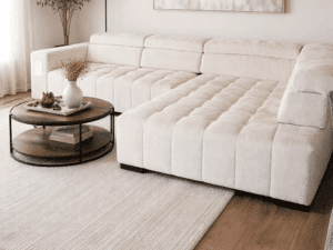 Canapé d'angle crème capitonné dans un salon moderne avec table basse ronde en bois et tapis beige texturé.