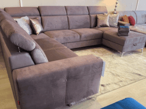 Canapé sectionnel en forme de U, velours mauve/gris-rose, avec repose-pieds et coussins décoratifs, sur plancher clair et tapis texturé.