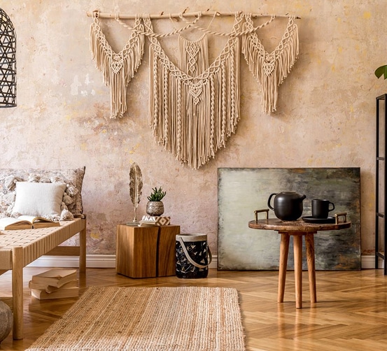 Décoration Bohème Chic : Macramé, Bois et Textures Naturelles Salon bohème chic avec mur texturé, tenture macramé beige, banc en bois tressé, tapis jute, table d'appoint en bois et théière noire.