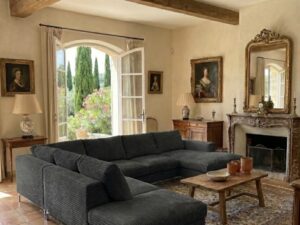 Salon aux poutres apparentes avec grand canapé d'angle gris, cheminée en pierre et jardin de cyprès en arrière-plan.