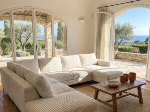 Salon méditerranéen lumineux avec canapé blanc en velours côtelé, poutres apparentes et terrasse vue mer.