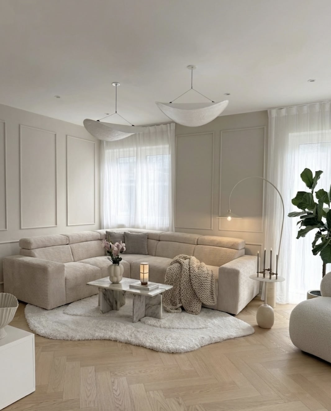 Salon minimaliste beige avec canapé d'angle côtelé, table en marbre, luminaires design et parquet en chevrons.