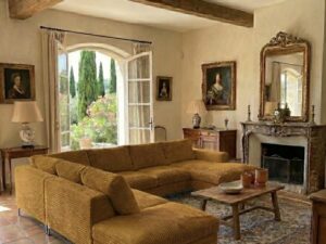 Salon provençal avec canapé d'angle ocre, poutres apparentes, cheminée ancienne et vue sur un jardin méditerranéen.