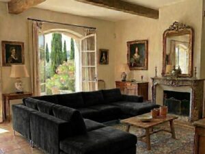 Salon rustique avec grand canapé d'angle noir, poutres en bois apparentes, carrelage en terre cuite et cheminée ancienne.