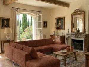 Salon rustique chic avec canapé en velours côtelé terracotta, poutres apparentes et vue sur un jardin provençal.