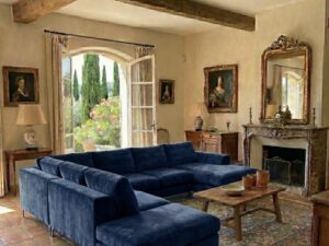 Salon rustique élégant avec canapé en velours bleu, poutres en bois et vue sur un jardin via des portes-fenêtres.