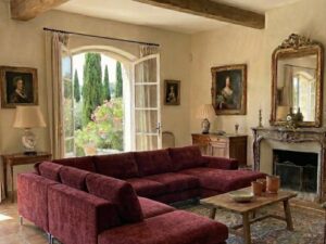 Salon élégant avec grand canapé bordeaux, poutres en bois et vue sur un jardin méditerranéen ensoleillé.