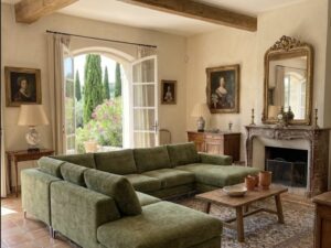 Salon rustique avec canapé d'angle vert, poutres en bois, cheminée en pierre et vue sur un jardin méditerranéen.
