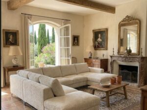 Salon rustique chic avec canapé d'angle, poutres en bois, cheminée ancienne et vue sur un jardin de cyprès.