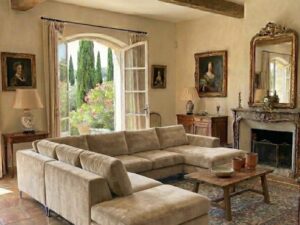 Salon rustique avec poutres en bois, grand canapé d'angle beige, tapis oriental et vue sur un jardin provençal luxuriant.