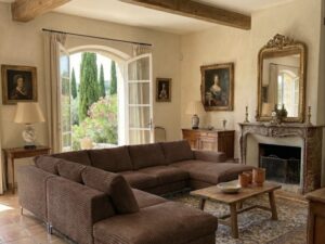 Salon chaleureux avec canapé en velours côtelé marron, poutres apparentes et vue sur un jardin méditerranéen.